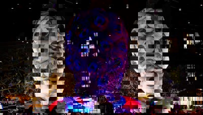 A large inflatable structure in the shape of a human head that is coloured purple and silver is covered in eyes looking outwards, with purple light all around.